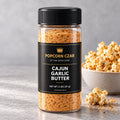 Product visualization of Popcorn Czar Cajun Garlic Butter in a round 2 oz shaker with a black-and-gold label beside seasoned popcorn. Actual batch shade may vary.