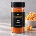 Product visualization of Popcorn Czar Chili Lime in a round 2 oz shaker with a black-and-gold label beside seasoned popcorn. Actual batch shade may vary.
