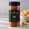 Product visualization of Popcorn Czar Smoky BBQ in a round 2 oz shaker with a black-and-gold label beside seasoned popcorn. Actual batch shade may vary.