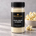 Product visualization of Popcorn Czar Sour Cream & Onion in a round 2 oz shaker with a black-and-gold label beside seasoned popcorn. Actual batch shade may vary.