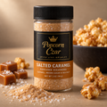 Popcorn Czar Salted Caramel popcorn seasoning in a 2 oz round shaker with an amber flavour band, shown with caramel pieces, flaky sea salt and a bowl of popcorn